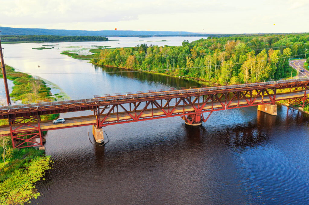 A red metal bridge spans a wide river surrounded by lush green trees, with a car crossing the bridge. This scenic spot is perfect for water activities in Superior, Wisconsin, set against distant forests and a partly cloudy sky.