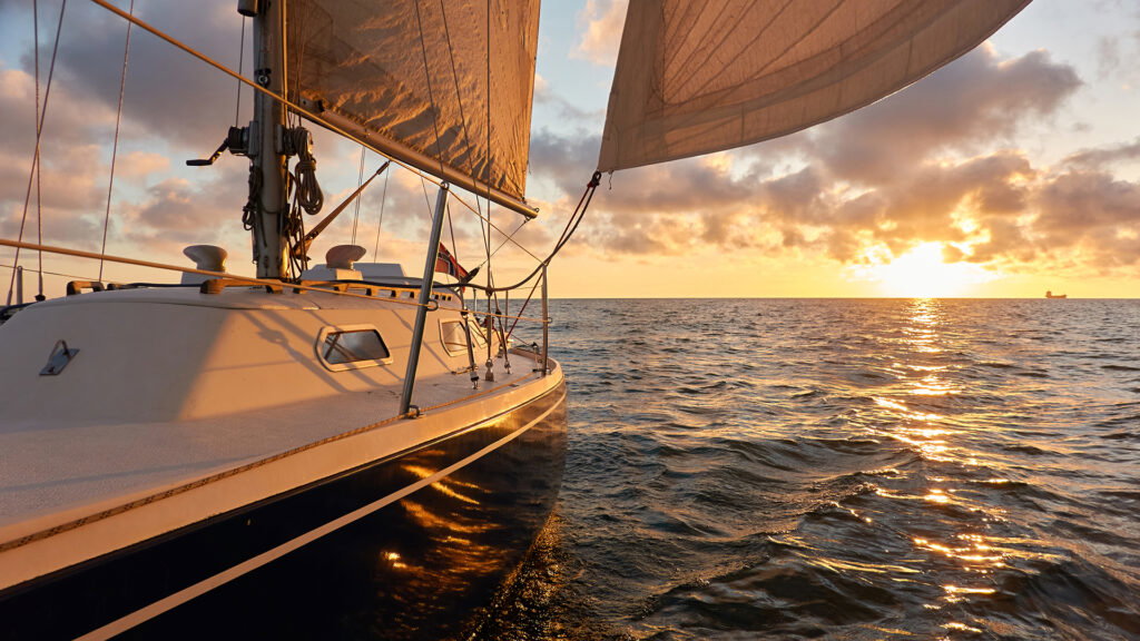 A sailboat glides on calm ocean water at sunset, with golden sunlight reflecting on the waves and partly cloudy skies—reminiscent of peaceful water activities in Superior Wisconsin.