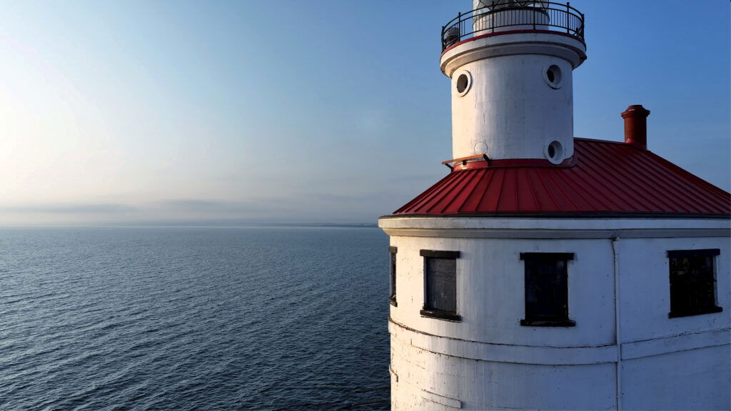 A white lighthouse with a red roof stands at the edge of calm blue water under a clear sky, bathed in sunlight—a perfect scene for a memorable Wisconsin family vacation.
