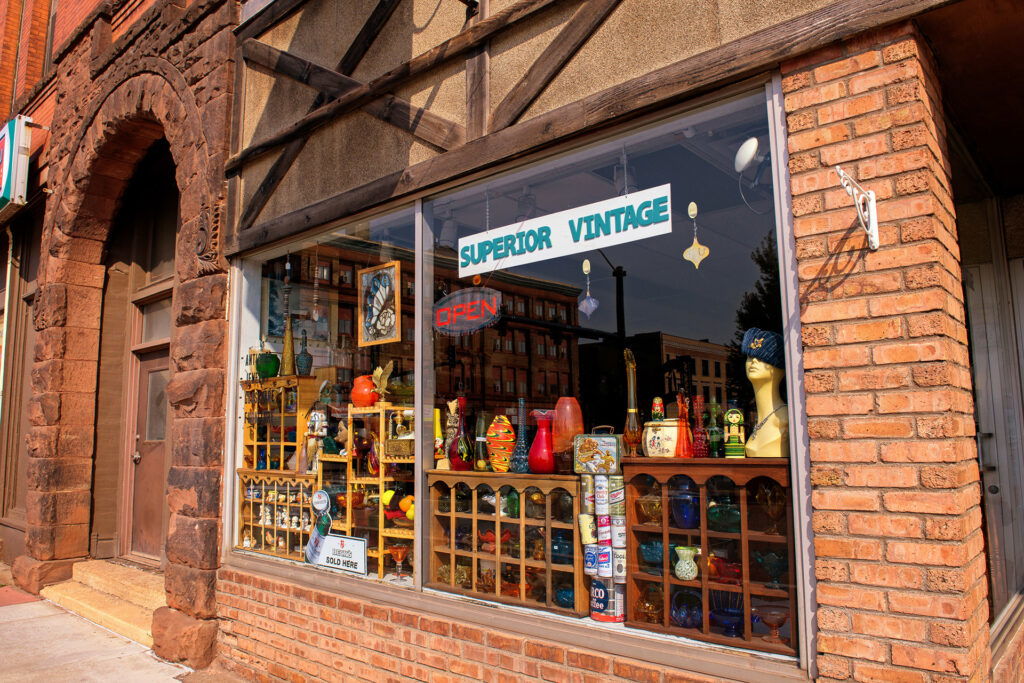 A brick storefront with large windows displaying vintage items, colorful glassware, and decorative objects beneath a "Superior Vintage" sign—a perfect stop for weekend getaways in Wisconsin. Reflections of nearby buildings shimmer in the glass.