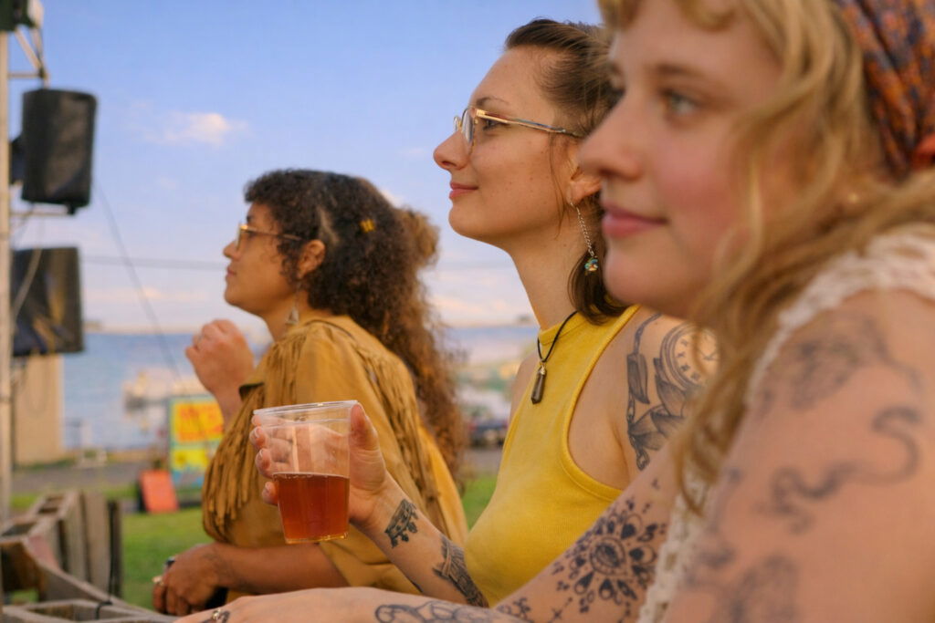 Three women with tattoos enjoy a sunny day near a waterfront during a Wisconsin Family Vacation. One holds a drink as they relax, watching an outdoor event by the stage, with water and green grass in the background.