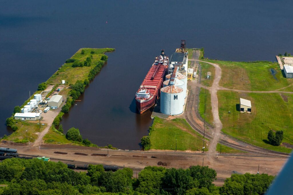 Aerial view of a large cargo ship docked next to silos and industrial buildings on a waterfront surrounded by greenery and rail tracks, not far from the renowned museums in Superior, Wisconsin.