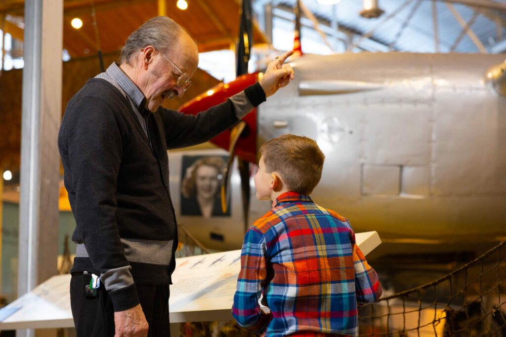 An older man smiles and points excitedly at an airplane in a museum, while a young boy in a colorful plaid shirt listens and looks at an informational display—one of the many fascinating things to do in Superior Wisconsin.