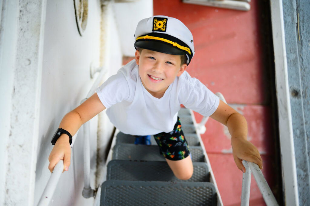 A smiling young boy wearing a white captain’s hat and t-shirt climbs up a metal staircase on a ship, looking up at the camera—capturing the excitement found at museums in Superior Wisconsin.