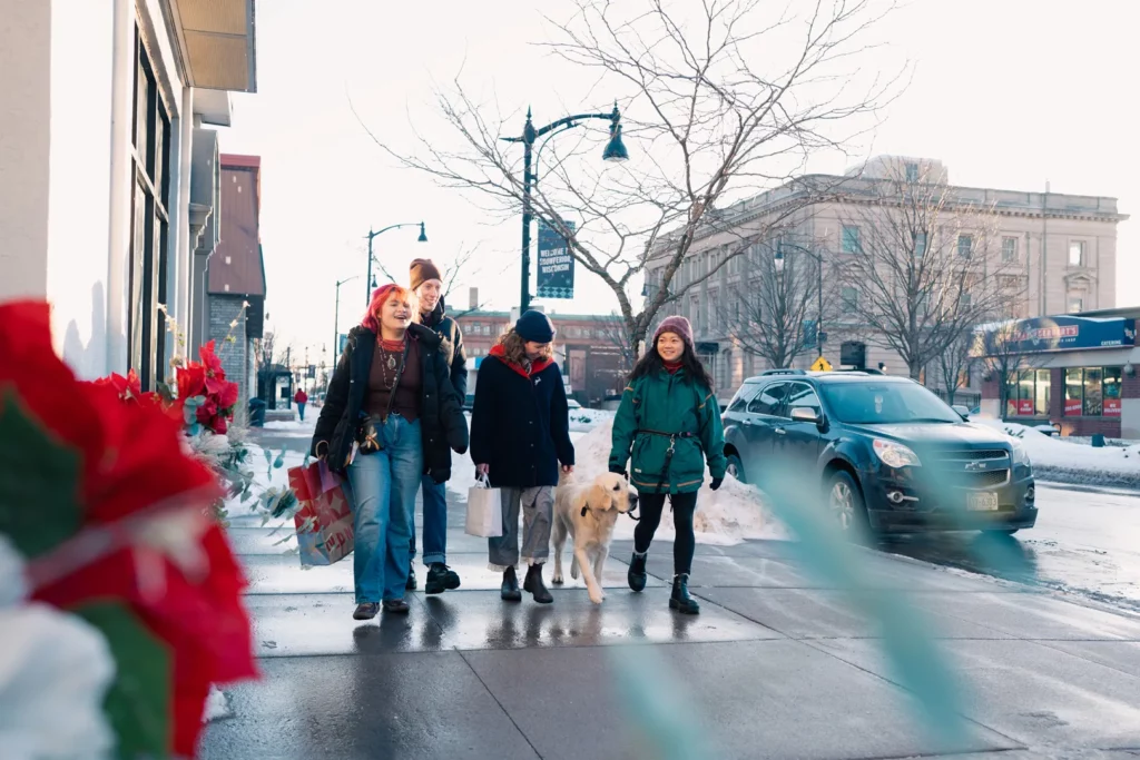 Four people walk on a snowy city sidewalk in Superior, Wisconsin with a dog; two carry shopping bags. Bundled in winter clothes and smiling, they pass red flowers. Cars and buildings line the street—a perfect snapshot of things to do in Superior, Wisconsin.