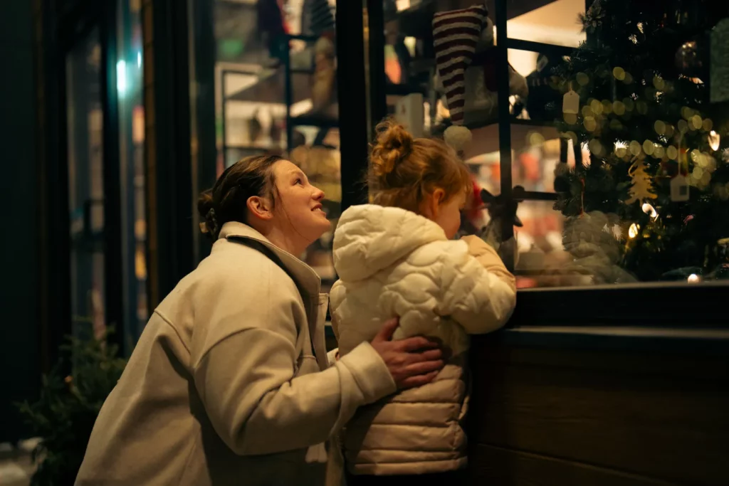 A woman and a young girl in warm coats look through a shop window decorated with Christmas ornaments and lights at night, appearing joyful and curious—capturing one of the festive things to do in Superior Wisconsin.