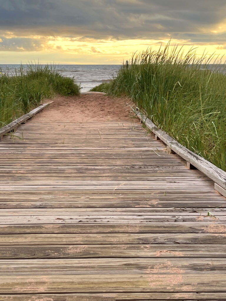 A wooden boardwalk leads through tall grass to a sandy beach, with waves and a dramatic, cloudy sunset sky in the background—perfect for memorable Wisconsin family vacations.