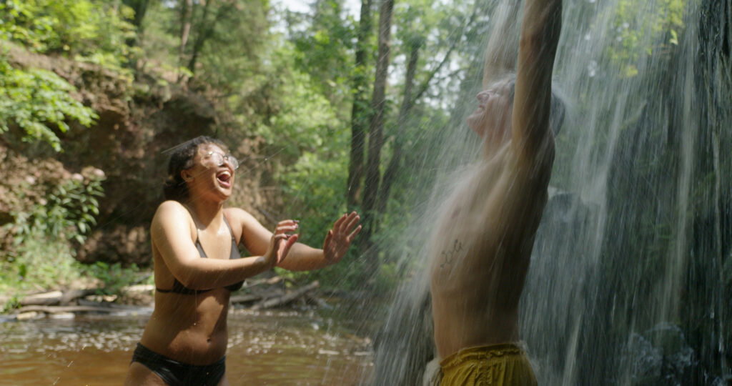 Two people in swimwear enjoy playing in a forest waterfall at one of the scenic parks in Superior, Wisconsin; one stands under the falling water with arms raised as sunlight filters through the trees and laughter fills the air.