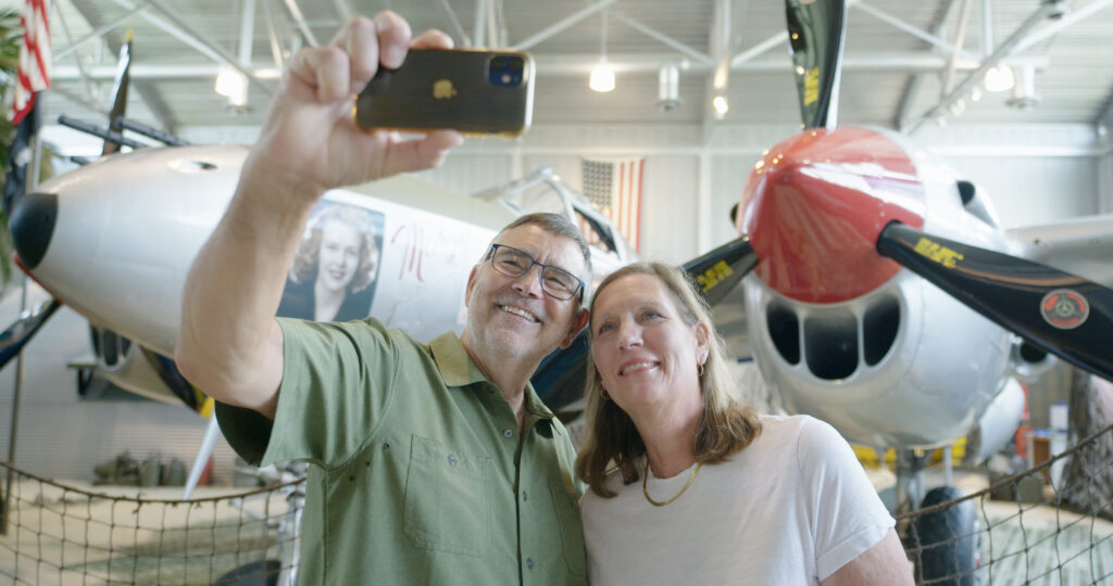 A man and woman smile as they take a selfie together inside an aviation museum in Superior, Wisconsin—one of the top things to do in Superior Wisconsin—standing before a vintage airplane with pin-up art and a red engine nose.