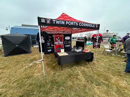 A red and black tent labeled "Twin Ports Cornhole" is set up outdoors on grass. Under the tent is a table with equipment, and several people stand nearby. The sky is overcast, and other tents and buildings are visible in the background.