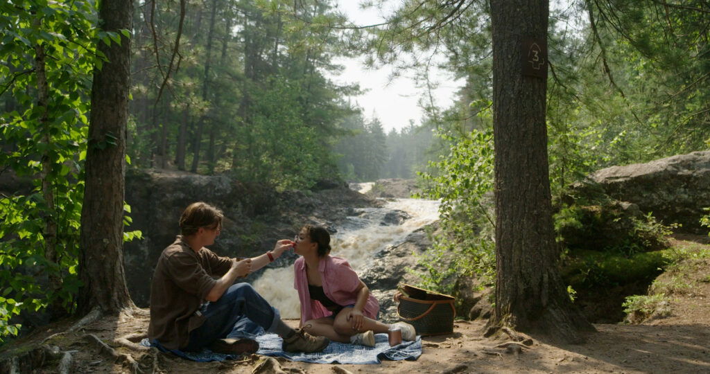 A couple sits on a blanket having a picnic in one of the scenic parks in Superior, Wisconsin, near a flowing river and waterfall. One person feeds the other as they smile, surrounded by trees, dappled sunlight, and a nearby picnic basket.
