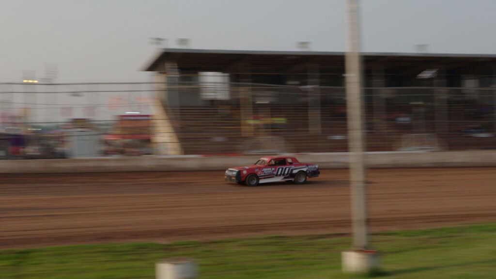 A red and white race car speeds along a dirt track in front of empty grandstands at a Wisconsin racetrack, with motion blur showing its movement—a thrilling scene for those seeking unique Wisconsin places to visit in summer. A pole is visible in the foreground.