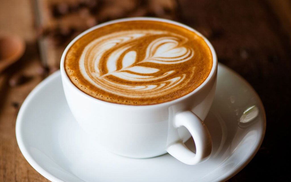 A white cup of coffee with intricate leaf-shaped latte art sits on a matching saucer atop a wooden table—a cozy scene you might find at some of the best places to eat in Superior WI.