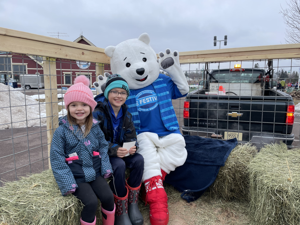 Two children in winter clothes sit on straw bales beside a person in a polar bear costume, all smiling during the Lake Superior Ice Festival. They are on a hay wagon with a pickup truck and snowy outdoor scene in the background.