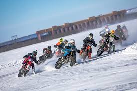 A group of motorcyclists, wearing colorful gear and helmets, race on a snow-covered track, kicking up snow as they navigate a turn under a clear blue sky.