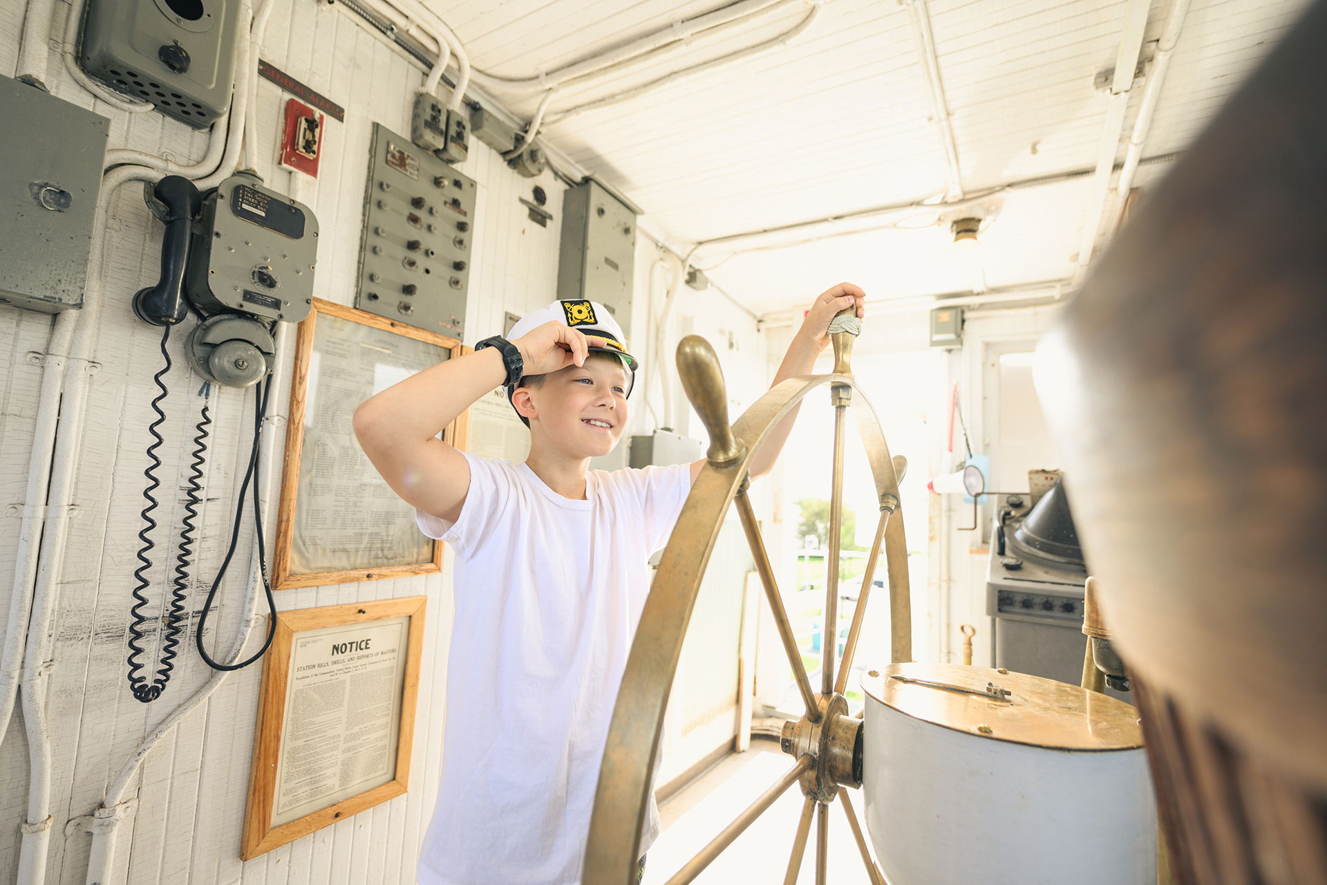 A smiling child wearing a captain's hat stands at a ship's helm, holding the wheel with one hand and touching the brim with the other, inside a bright vintage ship control room—perfect inspiration for Wisconsin places to visit in summer.