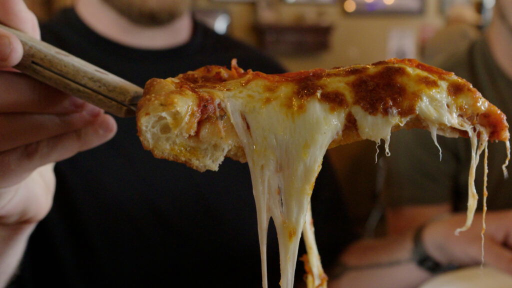 A person lifts a slice of cheesy pizza with a spatula, stretching melted cheese, while others sit in the background—capturing the flavor of Wisconsin places to visit in summer.