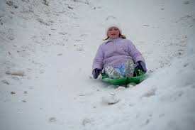 A young girl wearing a purple coat and white hat sleds down a snowy hill on a green sled, surrounded by snow.