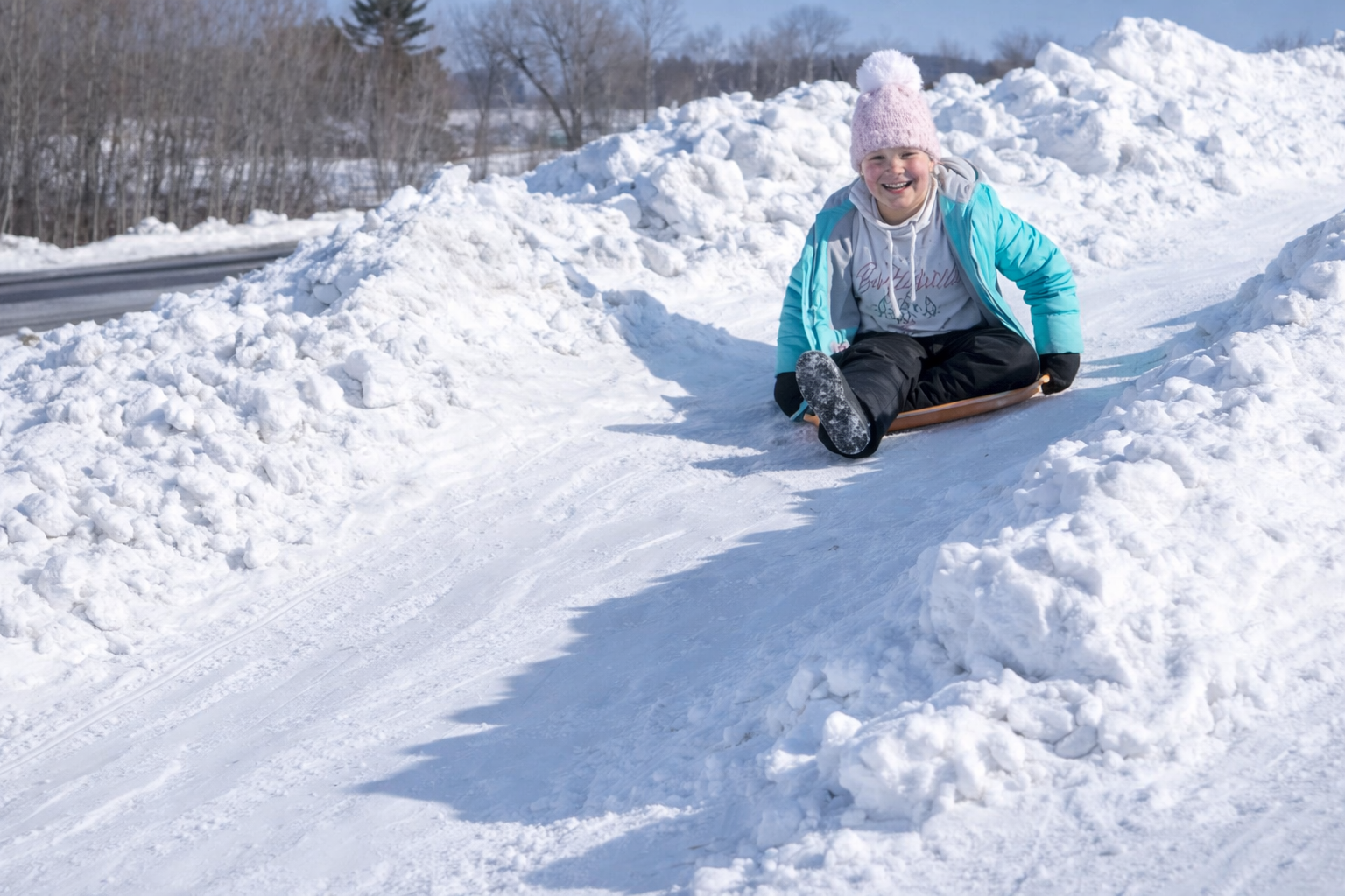 A smiling child in a blue jacket and pink hat sleds down a snowy hill surrounded by large snowbanks on a bright winter day at the Lake Superior Ice Festival.