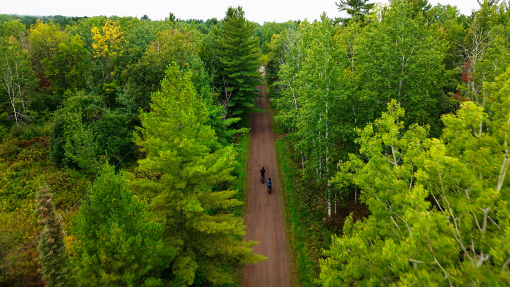 Aerial view of two people biking on a dirt trail surrounded by dense green forest with tall trees on both sides. This lush, vibrant scene highlights one of the best things to do in Superior Wisconsin for a peaceful outdoor adventure.