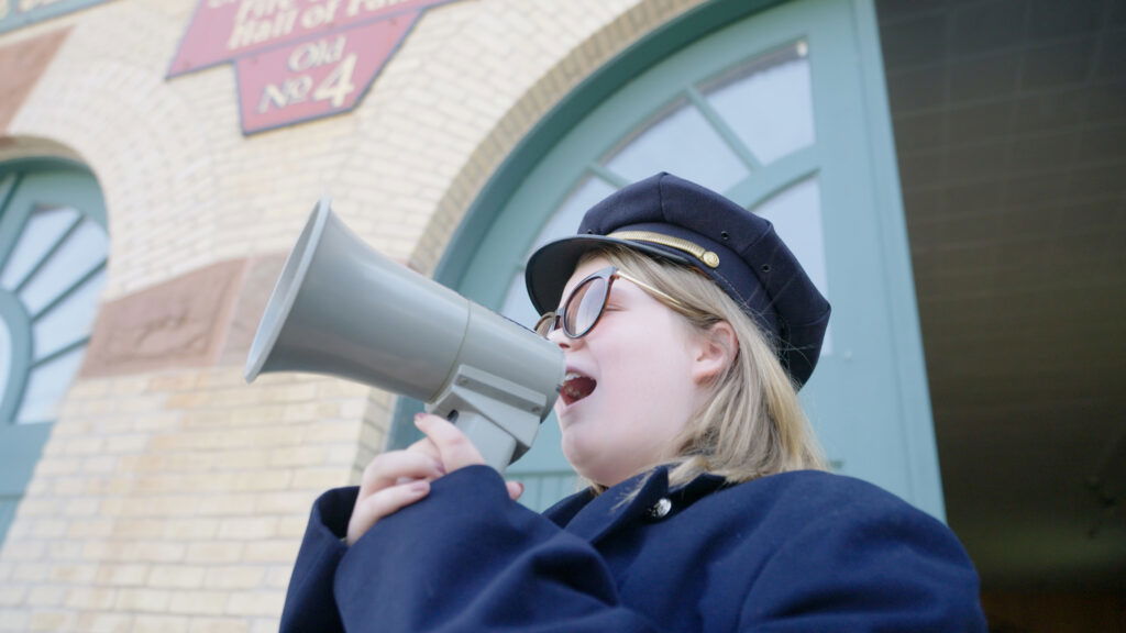 A person wearing a navy blue uniform and hat speaks into a gray megaphone outside a brick building, perhaps welcoming visitors arriving for weekend getaways in Wisconsin, with arched green doors and a red and gold sign above the entrance.