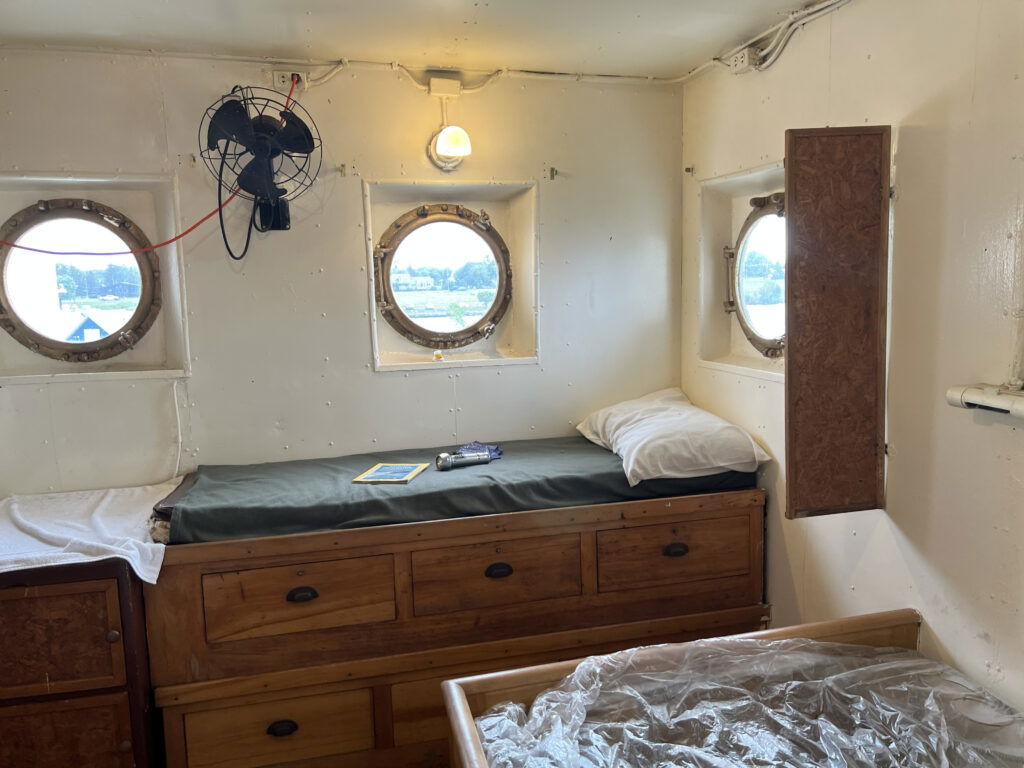 A cozy ship cabin ideal for Wisconsin family vacations, featuring three round portholes, a single bed with a green blanket and white pillow, wooden drawers beneath, a wall fan, and a book on the bed as light filters in from outside.