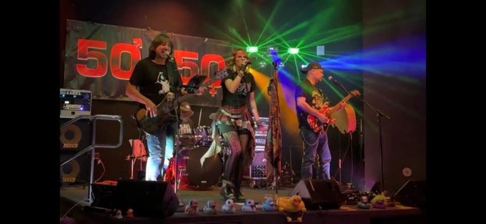 A rock band performs energetically onstage under colorful lights. Three musicians play guitar and bass, while a female vocalist sings at the center. The stage backdrop reads “50/50,” and toy ducks are lined up at the front edge of the stage.