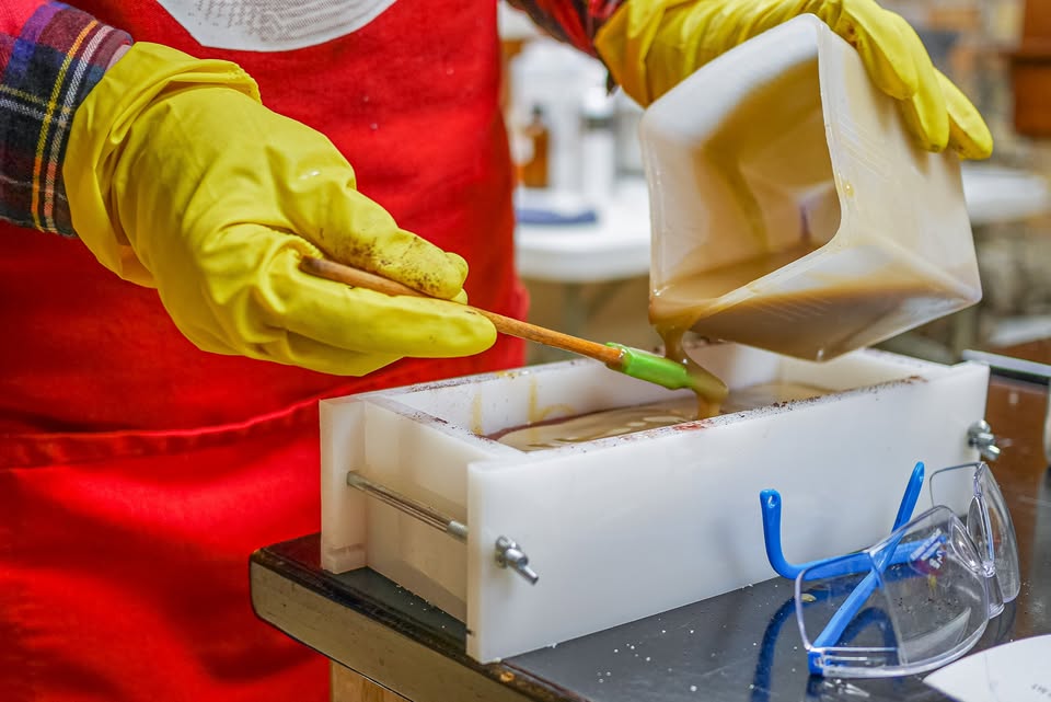 A person wearing yellow gloves and a red apron pours a brown liquid into a white mold, using a spatula to guide the flow. Protective glasses rest nearby on the table.