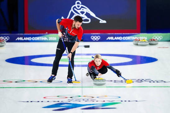 Two athletes compete in a curling match at the Winter Olympics. One is sliding a stone on the ice while the other sweeps in front of it. Olympic rings and the Milano Cortina 2026 logo are visible on the rink.