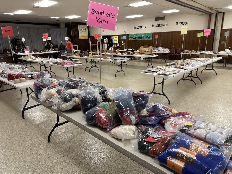 Tables in a large room display bags of yarn and craft supplies, each section labeled with signs such as "Synthetic Yarn." People are browsing in the background. The atmosphere is organized and inviting.