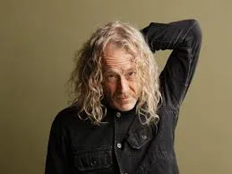 A man with long, wavy blond hair wearing a black button-up shirt stands against a plain olive green background, with one arm raised and his hand resting on the back of his head.