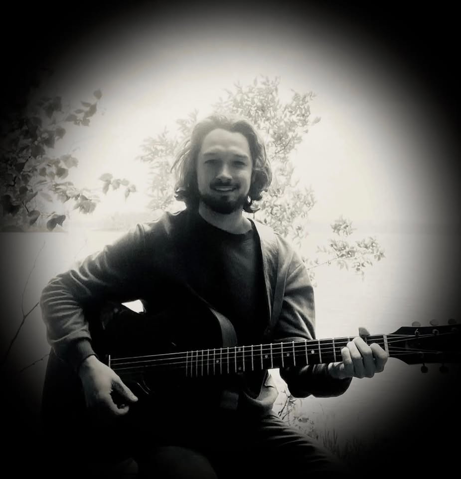 A person with long hair sits outdoors, playing an acoustic guitar. The image is in black and white with a vignette effect, creating a soft, glowing border around the subject. Trees and water are visible in the background.