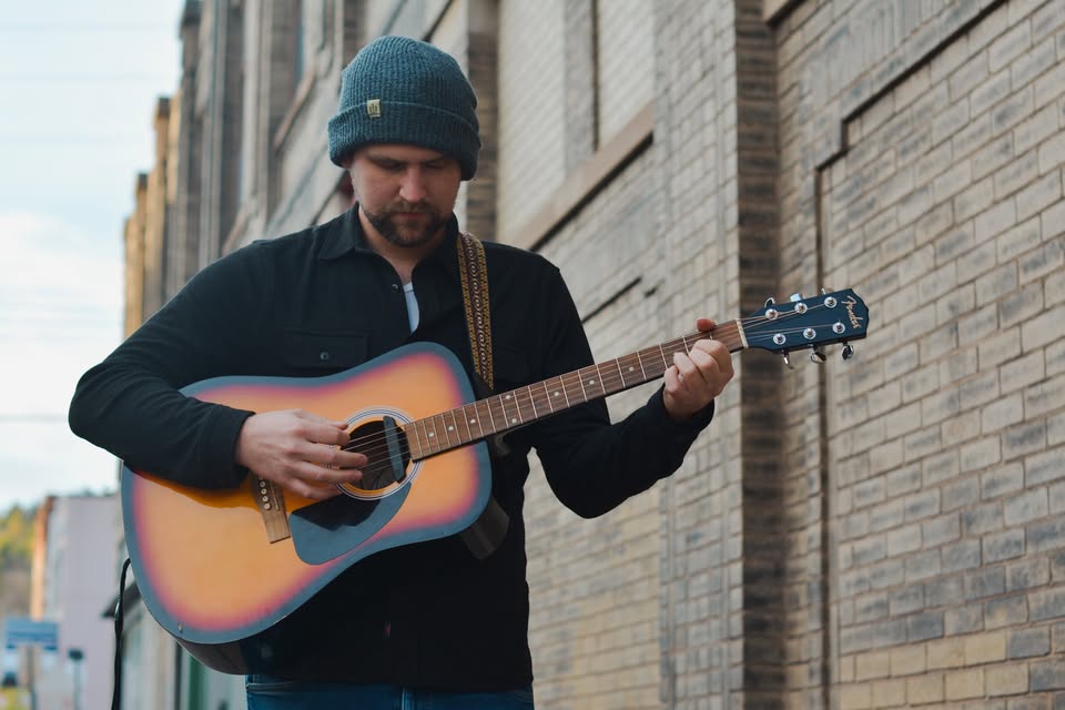 A man wearing a gray beanie and black jacket plays an acoustic guitar while standing next to a brick wall outdoors.