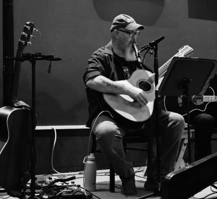 A bearded man in a cap sits on stage playing an acoustic guitar and singing into a microphone. Guitar cases and equipment are nearby. The image is in black and white.