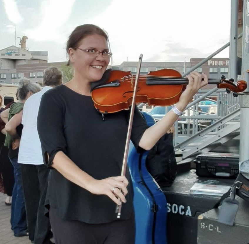 A woman wearing glasses and a black top smiles while playing a violin outdoors near a pier, with people and metal bleachers in the background.