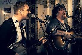 Two musicians passionately perform indoors; one sings and plays a banjo while the other sings and plays an acoustic guitar. A microphone and Irish-themed decor are visible in the background.
