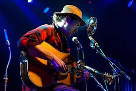 A musician wearing a hat and plaid shirt plays an acoustic guitar and sings into a vintage-style microphone on a dimly lit stage with blue and purple lights.