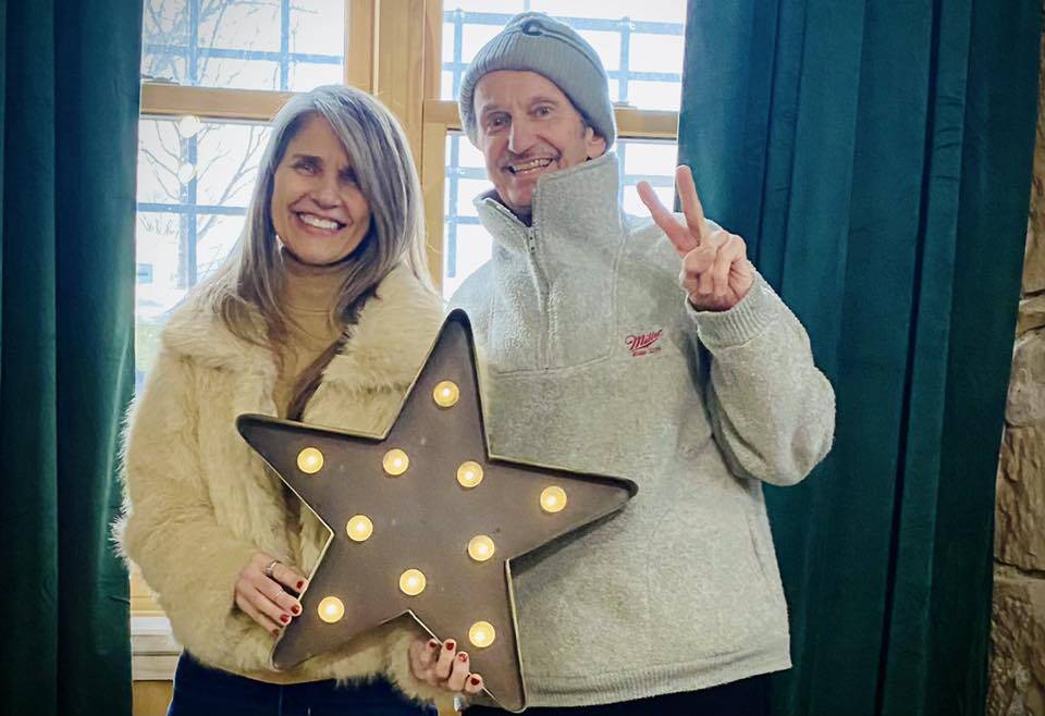 Two people stand indoors smiling. The woman on the left holds a metal star with lights, and the man on the right, wearing a beanie and gray sweater, makes a peace sign. Green curtains and a window are in the background.