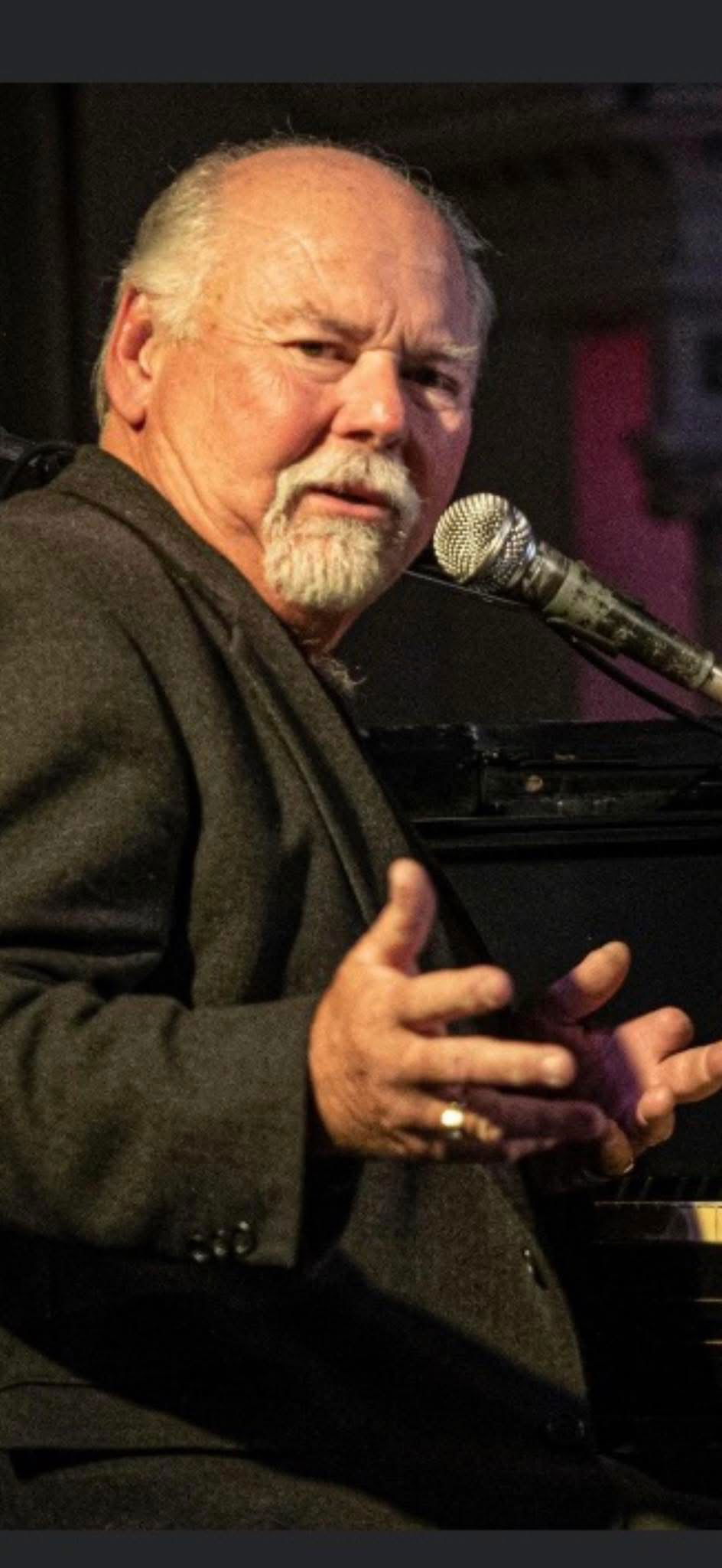 An older man with a mustache and goatee sits at a piano, gesturing with one hand while speaking or singing into a microphone. He wears a dark suit and looks intently at something off-camera.