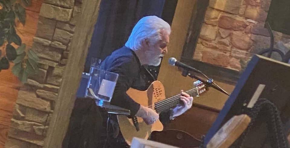 An older man with white hair and a beard plays an acoustic guitar and sings into a microphone at an indoor venue with stone walls and warm lighting.