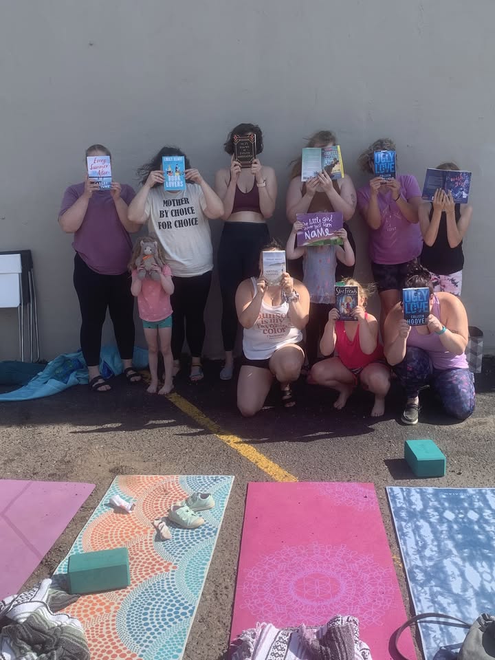 A group of nine people and one child pose outside against a wall, each holding a book in front of their face. Yoga mats and blocks are laid out on the ground in front of them.