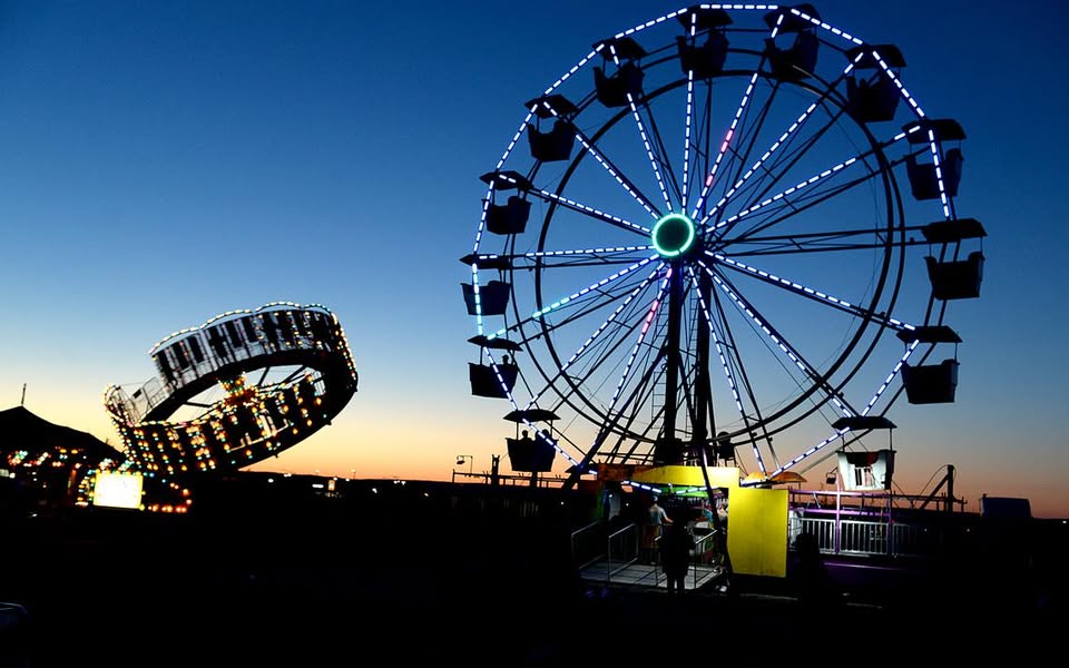 A brightly lit ferris wheel and a spinning amusement park ride glow against a deep blue sunset sky, with silhouettes of people and carnival booths in the foreground.