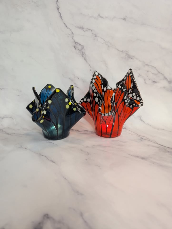 Two glass candle holders shaped like folded napkins, one with a blue and yellow butterfly wing pattern and the other with an orange, black, and white monarch butterfly wing pattern, on a marble surface.