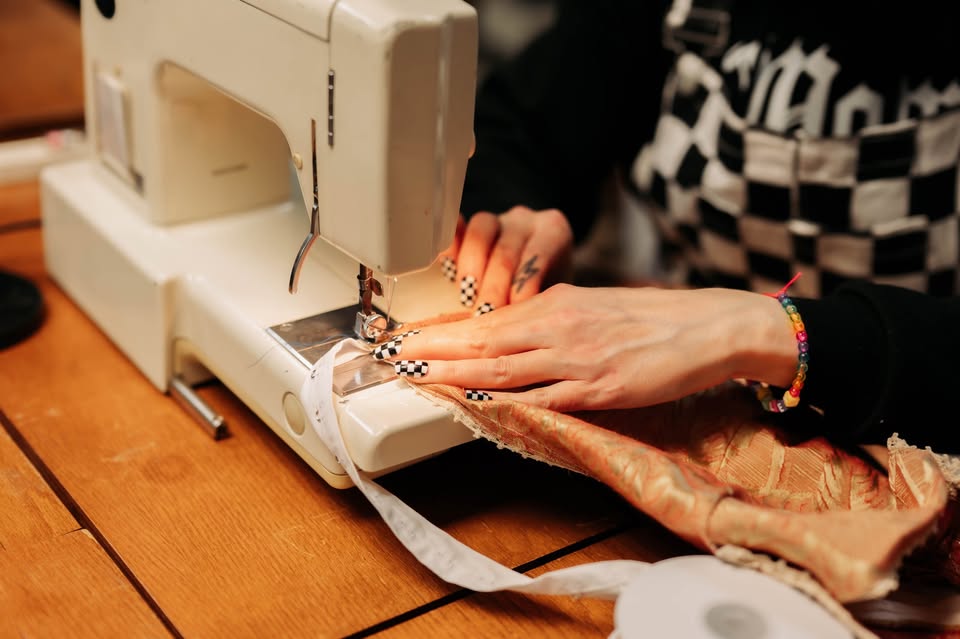 A person with checkered nails uses a sewing machine to stitch gold-colored fabric on a wooden table, wearing a black-and-white checkered apron and a colorful beaded bracelet.
