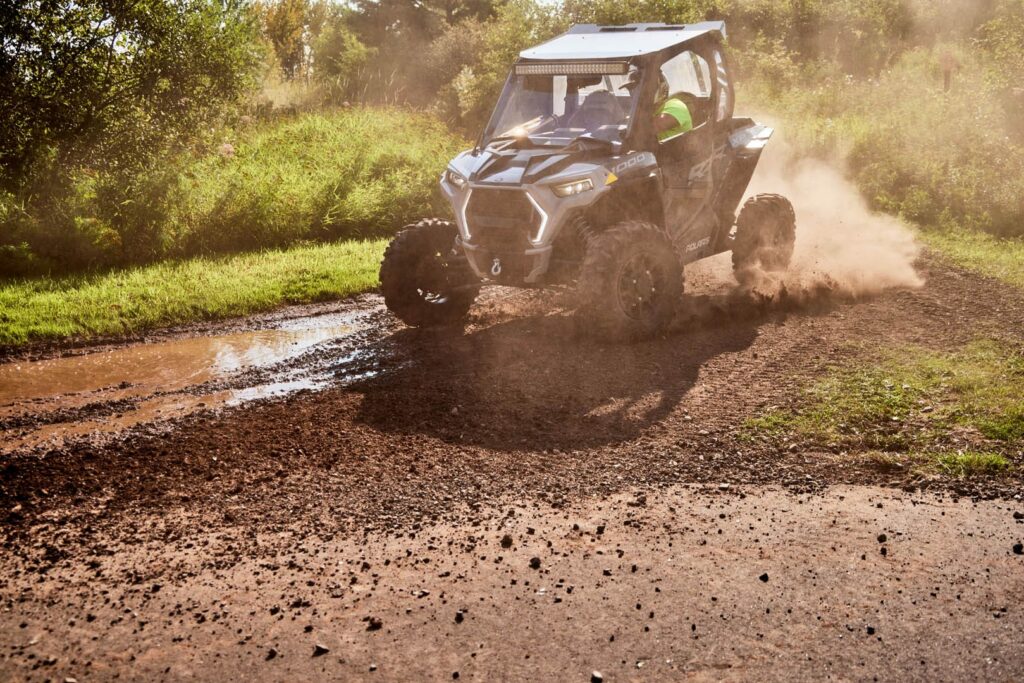 An off-road utility vehicle kicks up dirt and dust as it speeds along a muddy, gravel path—much like riders exploring Wisconsin snowmobile trails—surrounded by grass and trees on a sunny day.