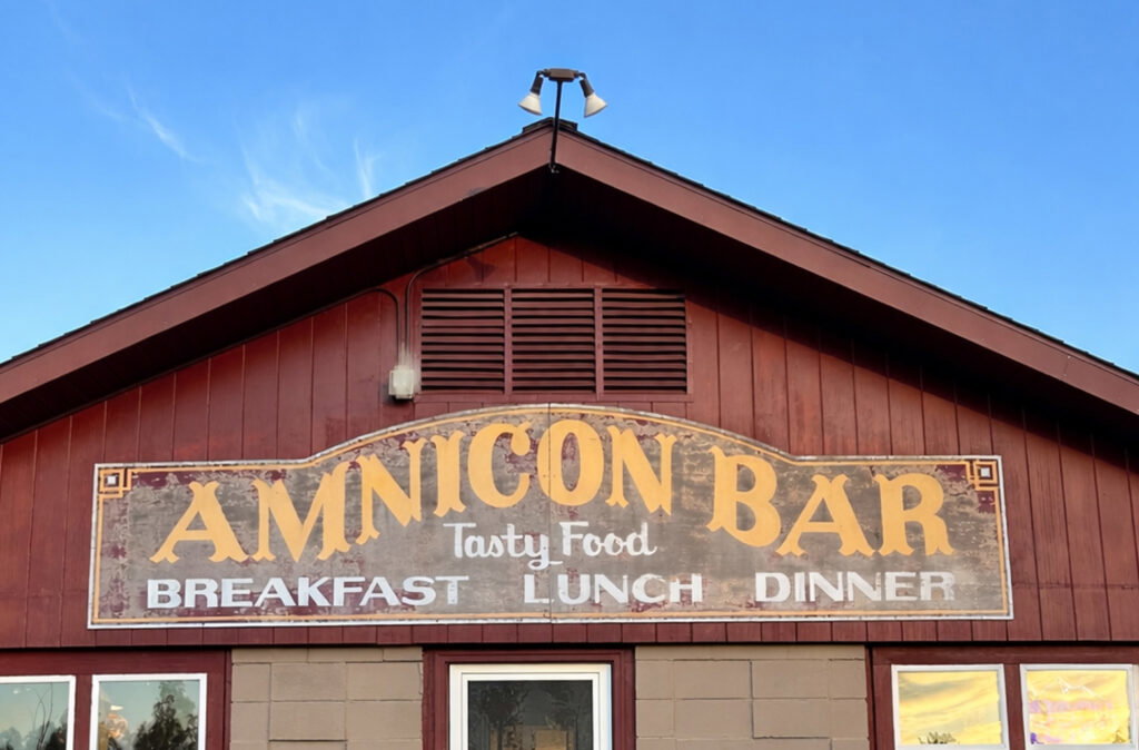 A building with a peaked roof and red siding features large windows advertising breakfast, lunch, and dinner. Overlaid is a semi-transparent graphic of a scale and book—perfect for travelers exploring nearby Wisconsin snowmobile trails.
