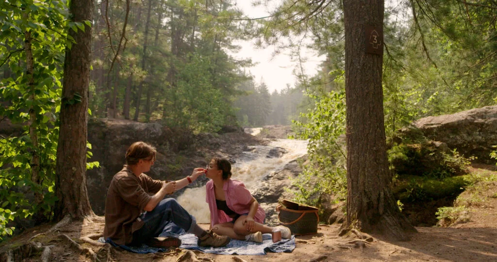 Two people sit on a blanket in a forest near a waterfall, sharing food. Enjoying a picnic on a sunny day, they’re surrounded by trees and nature—a perfect scene for those seeking Wisconsin places to visit in summer. A woven basket sits beside them.