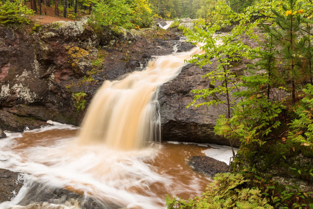A waterfall cascades over dark rocks into a brownish pool, surrounded by lush green trees and foliage—just steps from scenic Wisconsin snowmobile trails. The calm, natural scene features water flowing gently downstream through the forest.