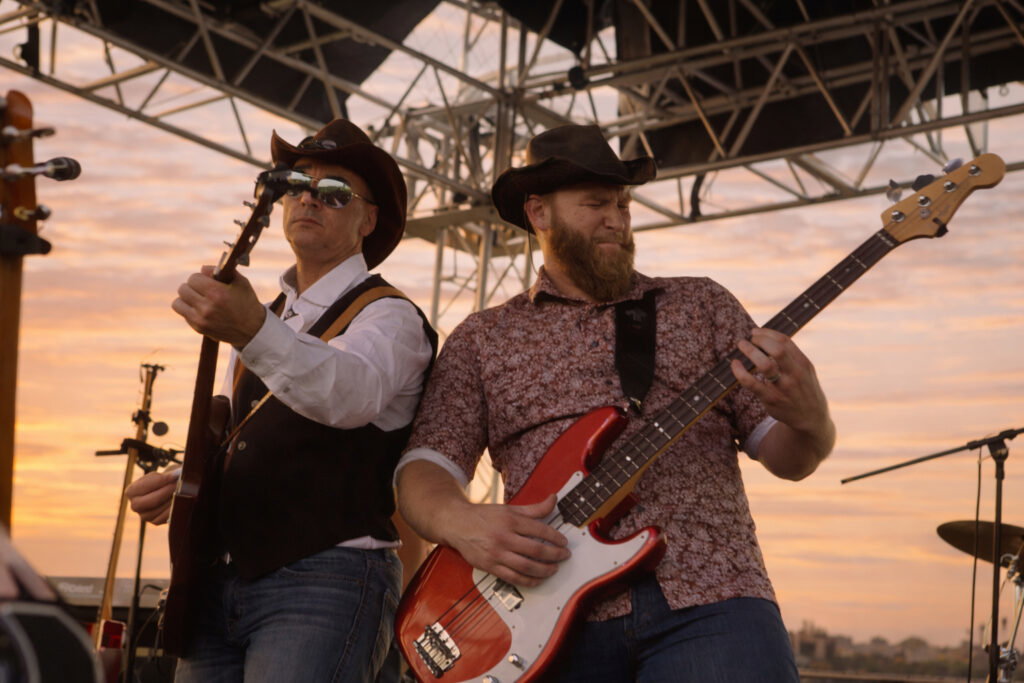 Two men in cowboy hats play electric guitars on an outdoor stage at sunset, embodying the lively music scene that makes Wisconsin places to visit in summer so memorable. Metal stage supports and a colorful sky form the perfect backdrop.