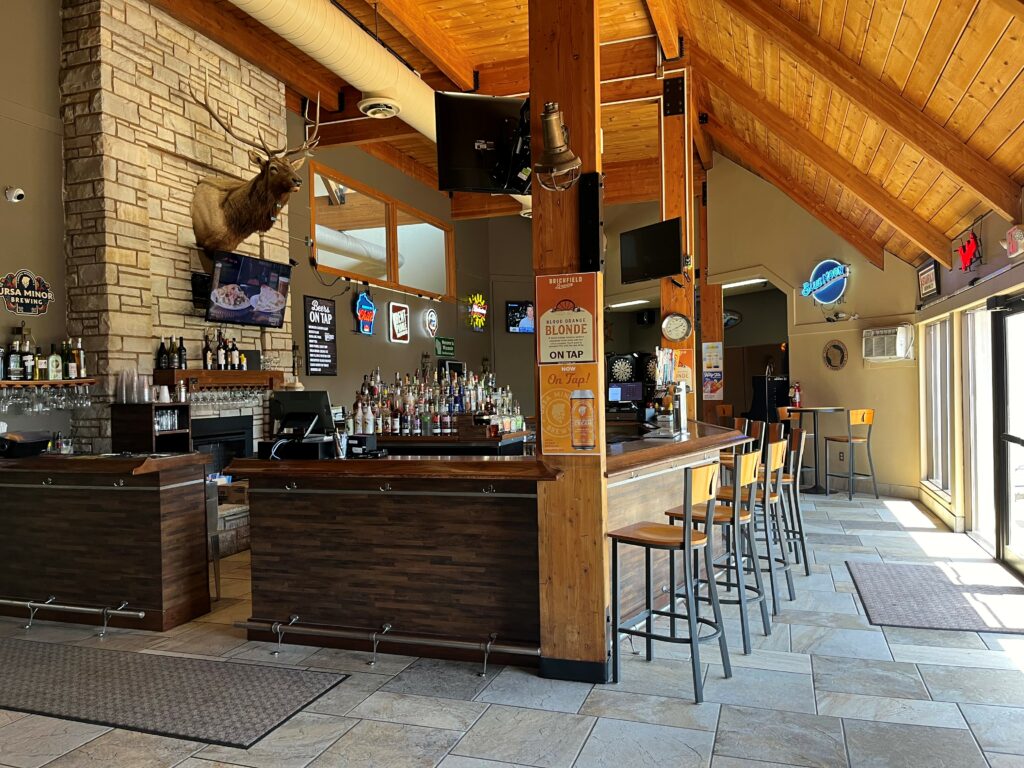 A rustic bar with wooden beams, stone fireplace, high ceiling, and mounted deer head invites travelers from nearby Wisconsin snowmobile trails to relax at the bar counter amid neon signs, liquor bottles, and large windows streaming in natural light.